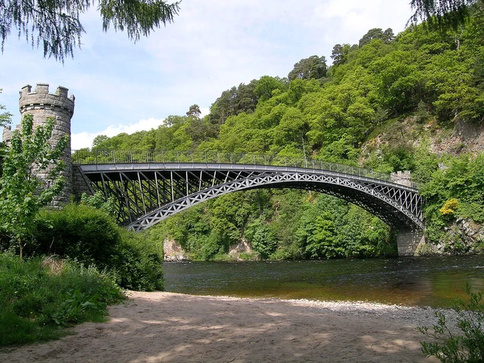 Craigellachie Bridge. The image shows the sandy shoreline of a river in the foreground with a rocky far shore rising steeply and covered in trees. An arched metal bridge crosses the river with abutments that look like castles from a chess set. The bridge has a lattice construction and is well above the viewer. The scene is in sunlight.