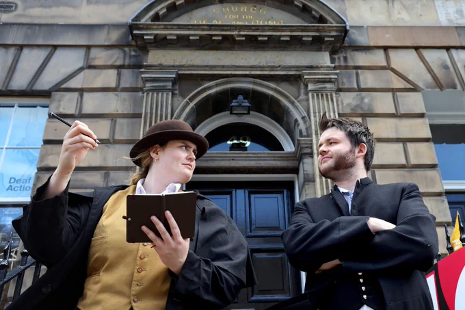 a woman and man stand outside a new town building, both dressed in victorian style clothing