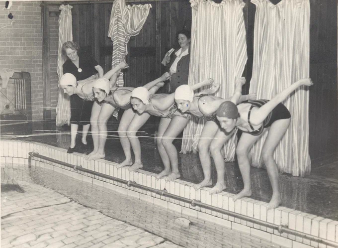 Black and white photograph of a row of five female students standing at the edge of a pool. They are wearing swimming costumes and caps. Their knees are bent and arms stretched behind them, poised to jump into the pool. Two fully dressed female members of staff look on. 