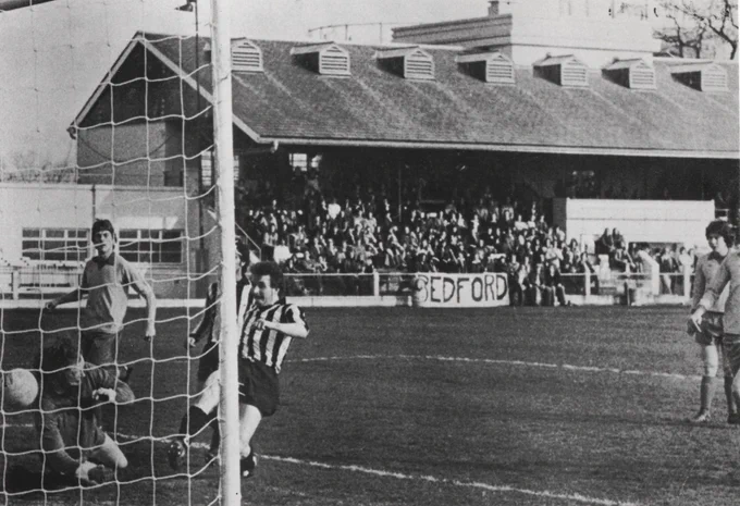 Black and white photograph of a men's football match. There is a crowd watching in the stand with a banner reading "Bedford". A player in a striped shirt is kicking a ball into a goal whilst the opposing goalkeeper is falling to the ground. Players in plain shirts look on.