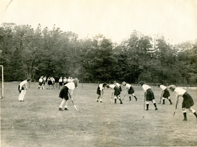 Black and white photograph of a women's hockey match on a field surrounded by trees. The female players are wearing knee-length skirts and holding hockey sticks.