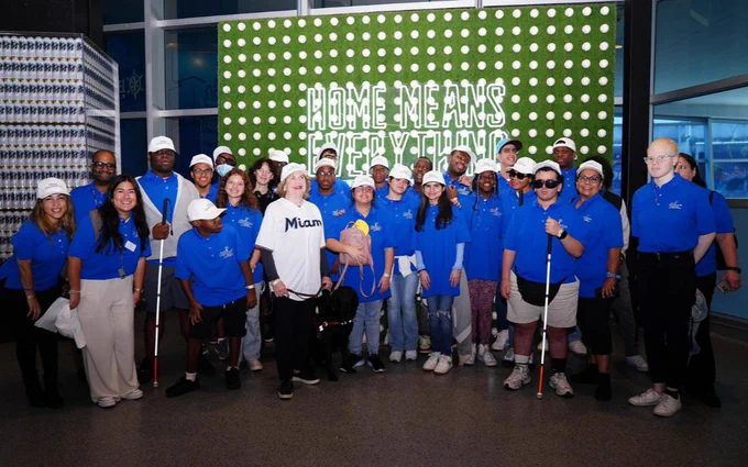 Group photo of 35 people smiling for the camera, in the photo everyone is wearing a blue Miami Lighthouse logo polo shirt, and 2 people are holding white canes. Virginia Jacko, CEO of Miami Lighthouse is in the forefront of the photo wearing a Miami Marlins logo jersey. 