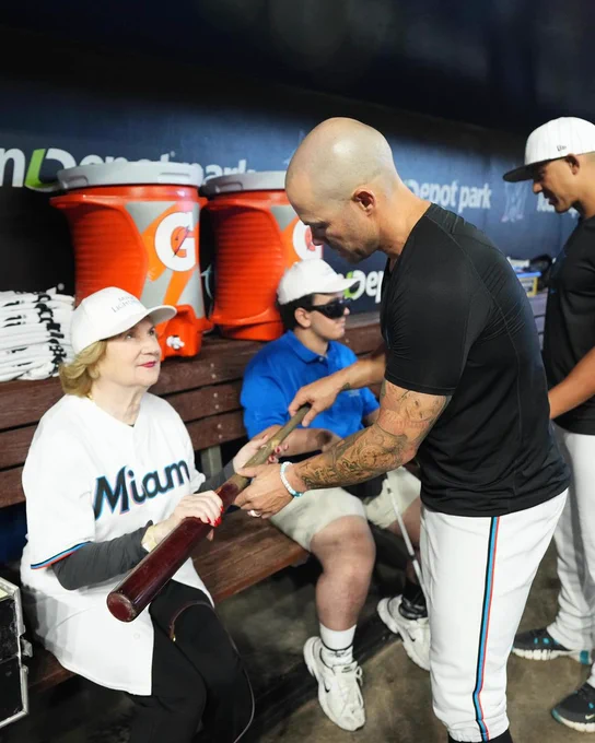 Photo: Virginia Jacko, CEO of Miami Lighthouse, holding a baseball bat with the guidance of a Miami Marlins professional baseball player explaining form.