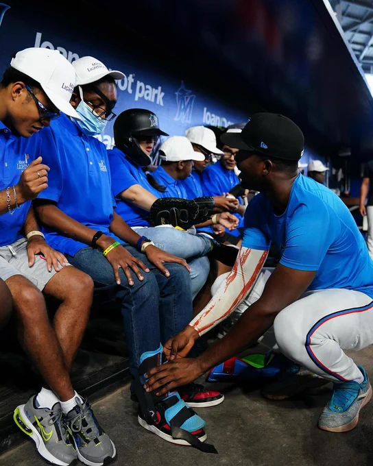 A photo showing a Miami Marlins professional baseball player helping a Miami Lighthouse teen student try on baseball equipment, with several classmates sitting nearby, wearing and exploring baseball equipment.