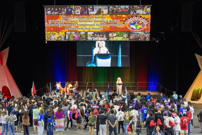 A crowd gathers before a stage at the Edmonton Expo Centre for the opening ceremony of the 2024 Alberta Indigenous Games. Red, yellow, blue and green horizontal lights colour the curtain behind the stage, which is flanked by two teepees. Hanging from the rafters overhead, a colourful banner reads "Alberta Indigenous Games. Canada's largest summer games and North America's Largest Indigenous Summer Games. Reclaiming our youth!"
