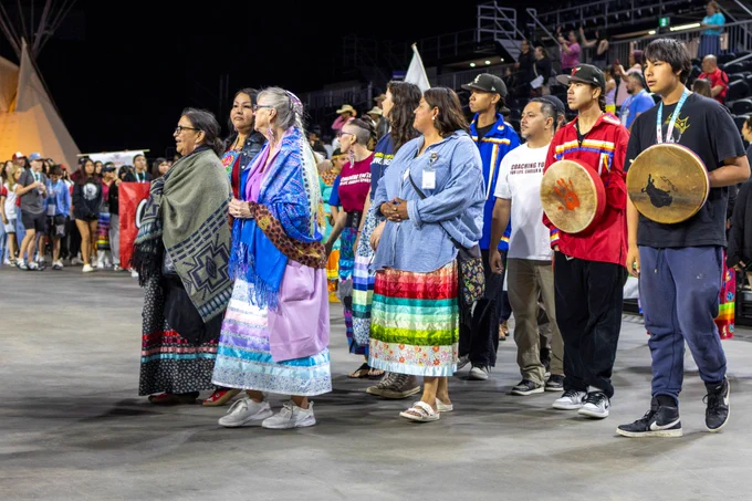 Participants in a grand entry dance in a circle around the Edmonton Expo Centre arena. Several of the women in the procession wear colourful ribbon skirts and shawls. Two men hold drums, one featuring a red palm print and the other a silhouette of a bison.