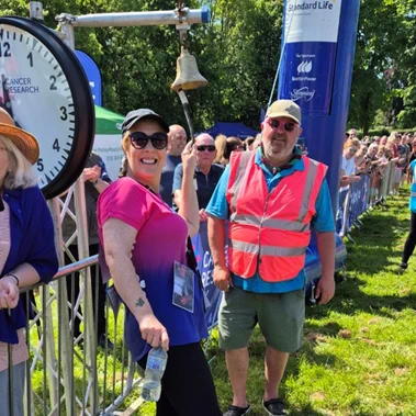Michelle smiling at the camera wearing a Race for Life t-shirt while ringing the bell at her local event.
