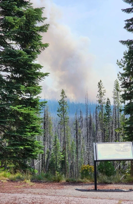 The Middle Fork Fire is visible in the distance, producing a large plume of smoke above the forest, viewed from a roadside with an informational signboard in the foreground.