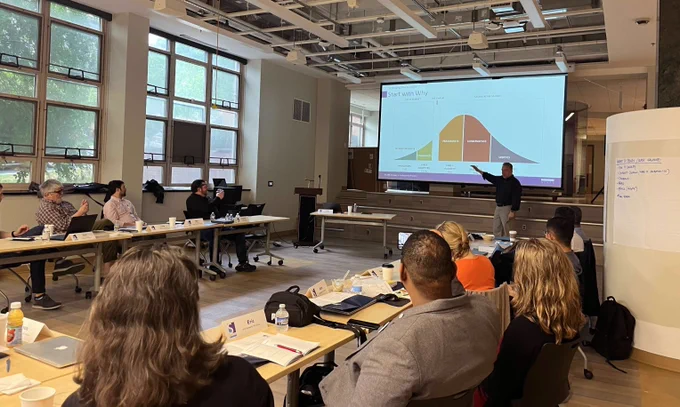 People sitting at desks watching someone present in front of a screen.