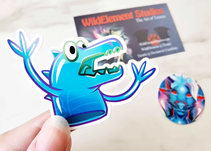 A photo of my hand holding a small sticker of a cartoon drawing of that 90s finger puppet dinosaur/monster toy. It's very cute. The artist's business card and pog are blurred out in the background.