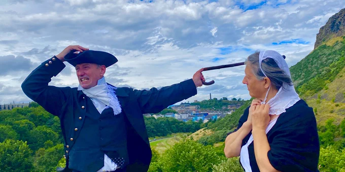 James Hutton, played by Billy Quain, and Jane Westhead as Isabella Hutton hold on to their hats with Salisbury Crags in the background.