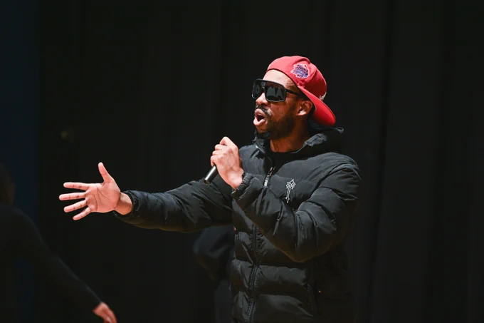 A black man in a puffer jacket, a red cap and dark glasses. He is rapping to an audience. 