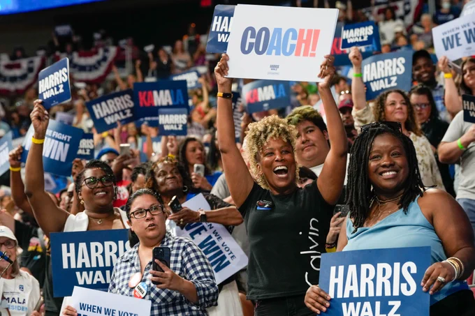 A crowd of Vice President Kamala Harris and Governor Tim Walz supporters hold signs that say "COACH!", "HARRIS WALZ", "KAMALA", and "WHEN WE VOTE, WE WIN".