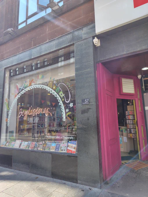 Bigglestones bookshopの店構え。ショーウィンドウにロゴとアーチ型の飾りと、それに沿うように草花、カメラ、犬、リス、ペイント。店内には書棚。入口はピンク