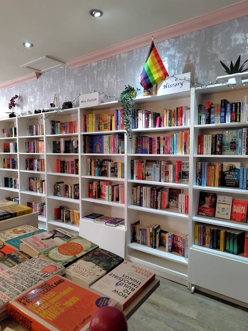 BigglestonesBookshop店内。ノンフィクションと歴史書の棚。白い本棚にたくさんの本が並んでいる。プライドフラッグが掲げられている。