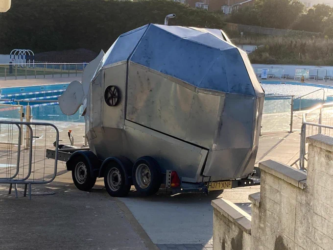 Big metal Whale on wheels in front of an outdoor swimming pool