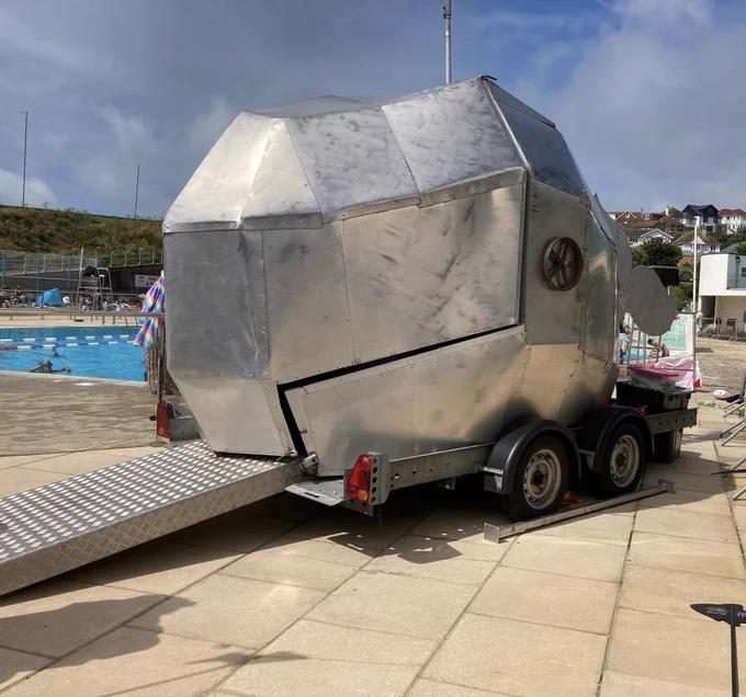 Big metal Whale on wheels with a ramp for a tongue in front of an outdoor swimming pool
