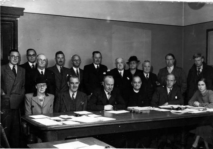 Lord Lindsay and the Advisory Committee 

Historical black and white photo of a group of 14 professionals, seated and standing around a large conference table