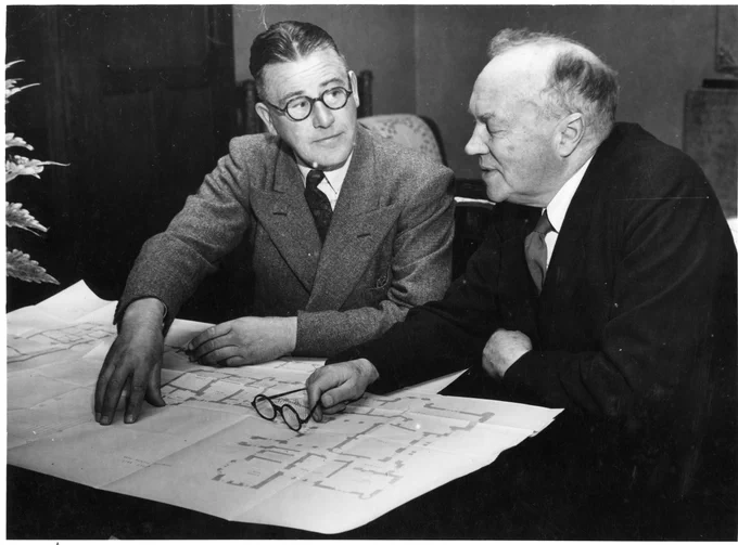 Two individuals, one being Lord Lindsay, are examining documents on a table, engaging in a focused discussion.