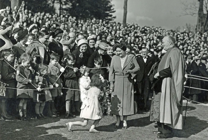 UCNS Opening Ceremony. The Queen Mother being greeted by a young child presenting flowers, with a crowd and Lord Lindsay in the background during a sunny outdoor event.