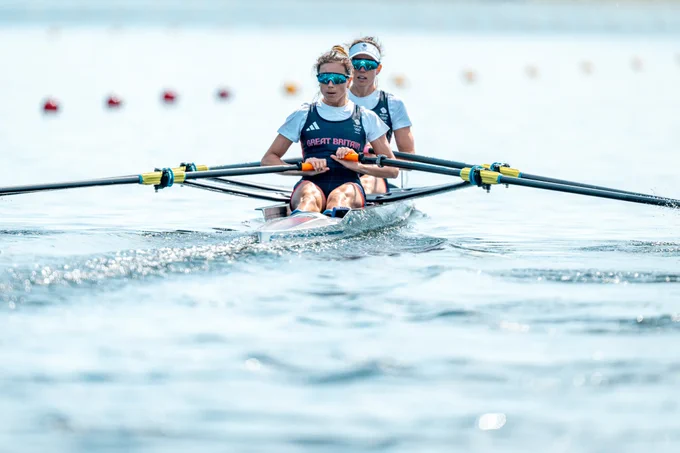 Imogen Grant and Emily Craig rowing in a double scull boat, wearing blue and white uniforms on a body of water marked with rowing lane buoys.