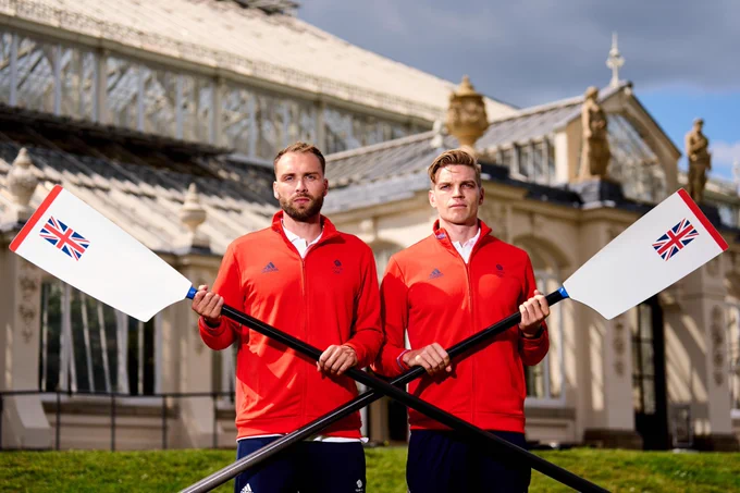 Ollie Wynne-Griffiths and Tom George in red fleeces and blue shorts holding a blade each in a cross over each other in front of Kew Gardens Temperate House.