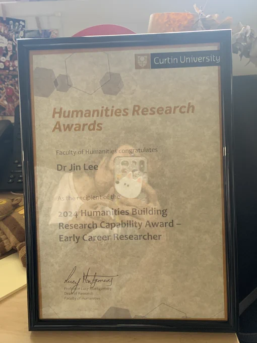 A photo of Dr Jin Lee's Curtin University Humanities Research Award: '2024 Humanities Building Research Capability Award – Early Career Researcher'. In the reflection of the award you can see Dr Jin Lee taking the photo with her phone.