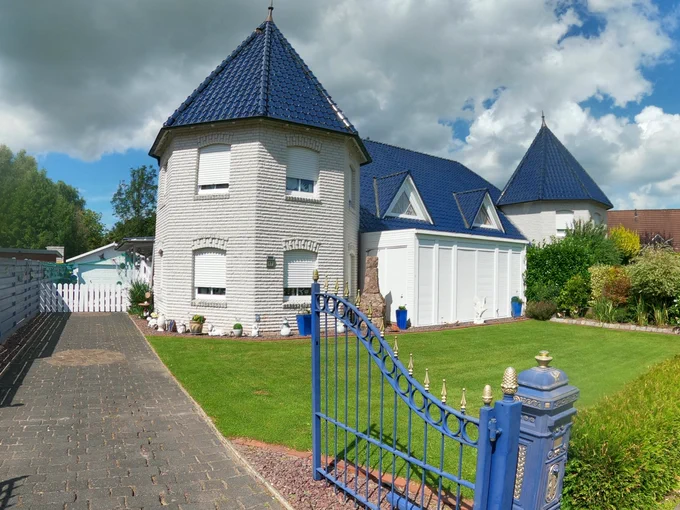 Ein Wohnhaus mit zwei Türmchen und großer Rasenfläche davor.  Ein Gartentor mit Auffahrt.
Das Einfamilienhaus ist doch recht hässlich. 