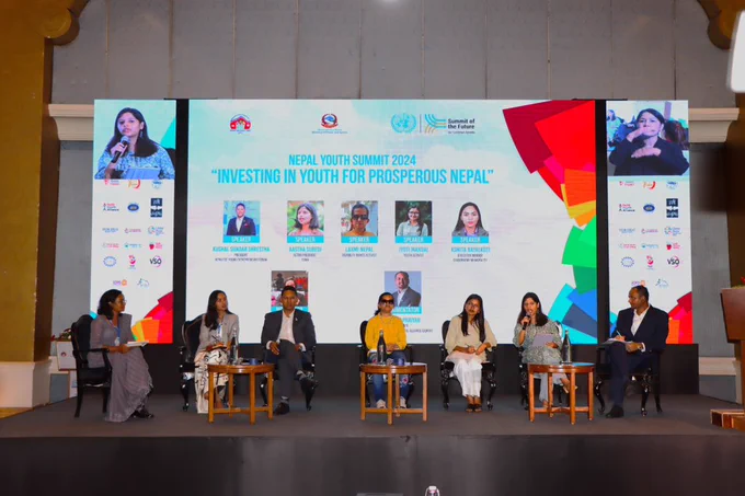 Panelists engage in a lively discussion at the Nepal Youth Summit 2024, held under the theme 'Investing in Youth for Prosperous Nepal.' The stage is set with six speakers seated in front of a large screen displaying the event’s title, speaker photos, and names. The backdrop includes logos of various organizations supporting the Summit of the Future.