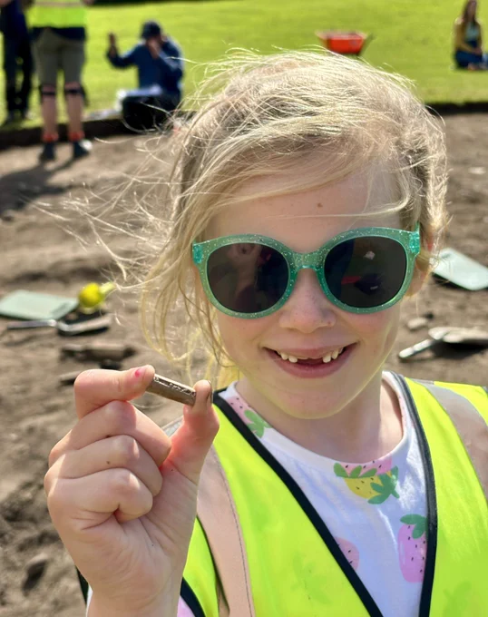 Photo of a girl in a high viz vest and sparkly green sunglasses holding a piece of a clay pipe with the archaeological trench in the background.