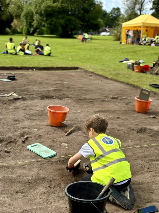 Photo showing the back of a young boy in a high viz vest who is troweling in a large archaeological trench.