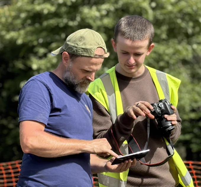 Photo of two men looking at a phone. The man on the right is wearing a high viz vest and holding a camera.