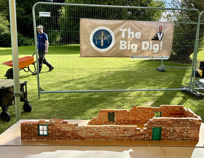Photo of a part-built brick model of a long, narrow WW2 building with a green door and window. Behind the model there is a large sign for the Big Dig and to the left an archaeologist pushing an orange wheelbarrow.