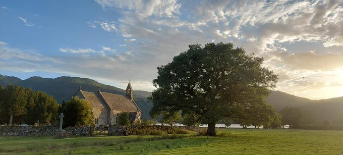 Small church of St Bega in the setting sun on the shore of Bassenthwaite Lake, oak in foreground, fells in background.