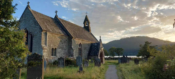Close up of small church of St Bega in the setting sun on the shore of Bassenthwaite Lake, oak in foreground, fells in background. Church surrounded by gravestones.