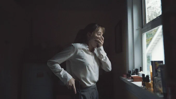 A woman in her late 20s (Maika Monroe) stands in a darkened room peering out the window in horror at something unseen. She covers her mortified expression with her left hand while reaching for a holstered gun on her waistband with her right hand. The daylight from the window softly illuminates the room.