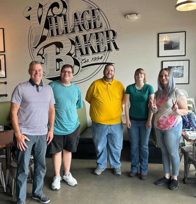 Five smiling auticon employees are wearing casual clothes and standing in a row in front of a wall with a large painting of the words Village Baker.