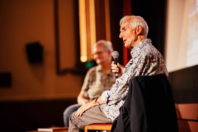 Peter Mitchell sat on HPPH stage and talking into mic. Blurred Martin Parr in background.