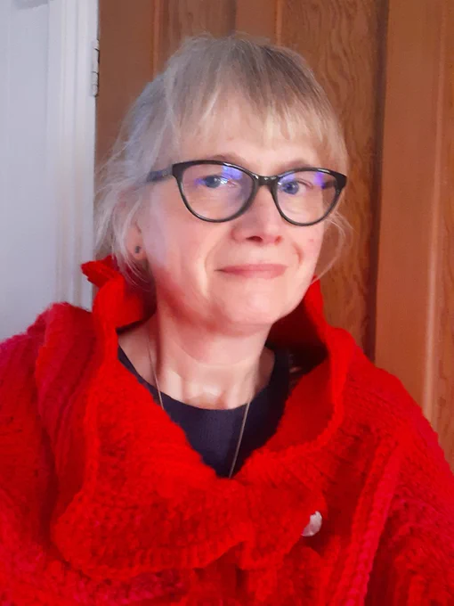A woman wearing glasses and a red knitted scarf standing indoors next to a wooden door.