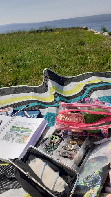Outdoor crafting scene with a sketchbook featuring a watercolor painting of a landscape, yarn, beads, and crafting supplies laid out on a colorful blanket, overlooking a sunny coastal view.