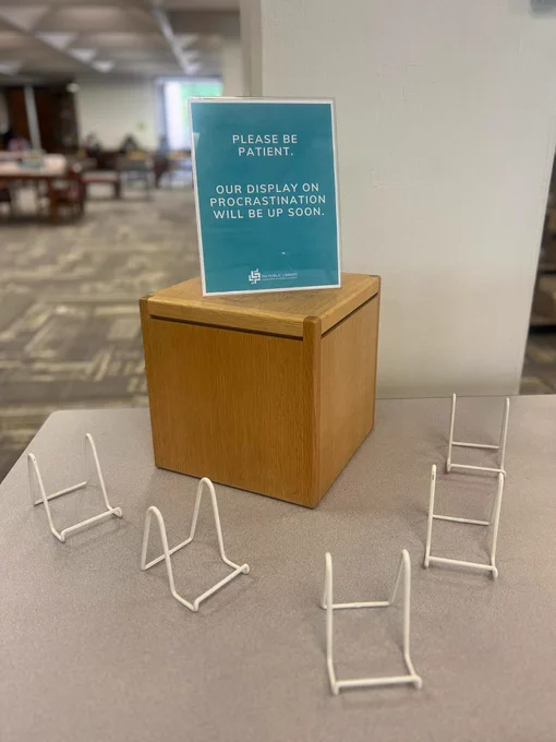 A photo of a book display with no books and a sign that reads “Please be patient. Our display on procrastination will be up soon.”