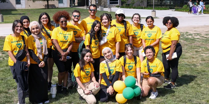 Group of students wearing yellow t-shirts smiles for a photo on a sunny day at a campus event, some holding green and gold balloons.