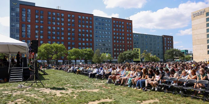 Large audience gathered on a grassy field for an outdoor event at Wayne State University, with a speaker addressing the crowd from a tent on the left.