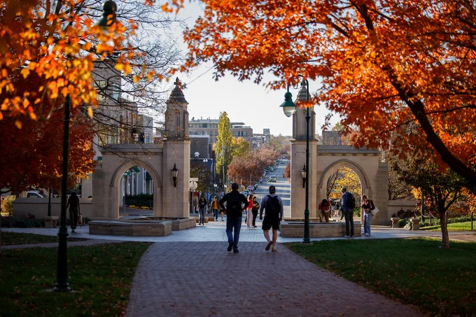 Two people walk on a brick path toward IU's iconic limestone Sample Gates, with Kirkwood Avenue and Bloomington's businesses just beyond.