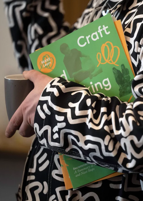 A person holding a mug and wearing a patterned jacket holds a copy of the MAKE Learn report tucked under one arm.