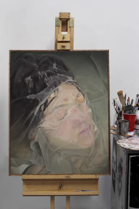 The painting presented on an easel in a studio space with a table and glasses filled with paint brushes on the right.