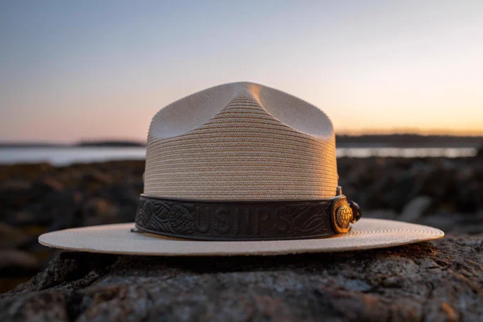 A close-up of a classic NPS ranger flat hat bathed in sunset light, resting on a rocky surface near the Atlantic Ocean.