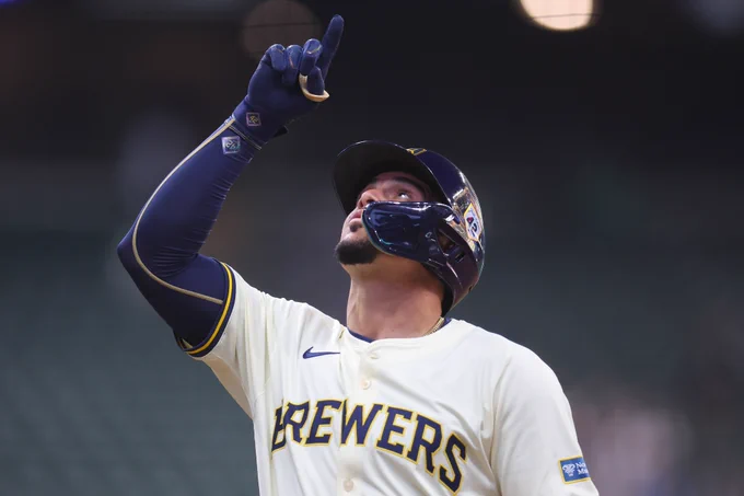 Willy Adames celebrates and points to the sky. He is wearing a cream Milwaukee Brewers jersey with navy and yellow lettering and Brewers batting helmet.
