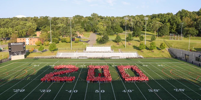 Aerial view of a large group of people forming the number "200" on a football field, surrounded by trees and buildings, under a clear sky.