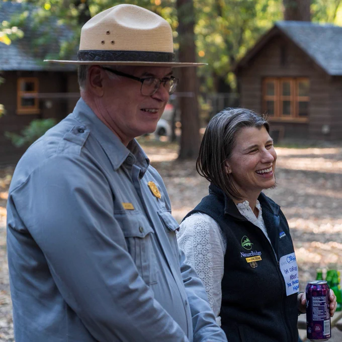 An NPS ranger wearing the NPS flat-brimmed hat and ranger uniform smiles and stands next to a smiling NatureBridge staff member wearing a black vest with the NatureBridge logo.